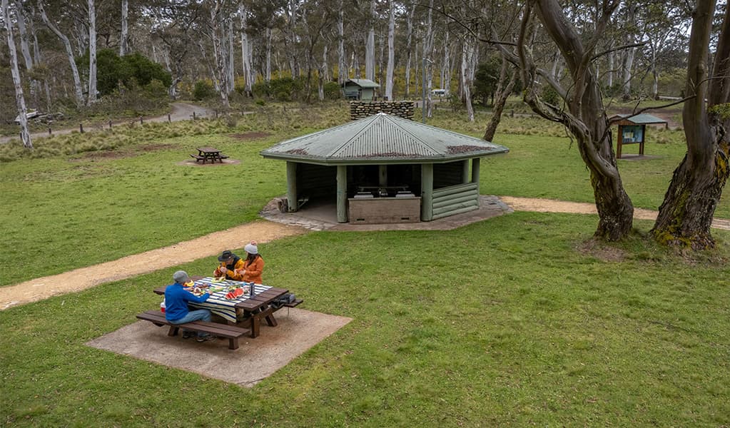 2 people eating at a picnic table near barbecue facilities under a covered pavilion in Polblue campground and picnic area. Credit: John Spencer/DCCEEW © DCCEEW