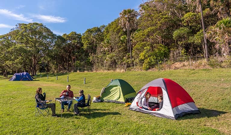 People sitting at a camping table drinking tea, while their kids play in their tent. The Ruins campground in Booti Booti National Park. Photo credit: John Spencer © DPIE