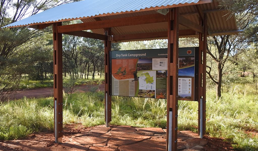 Photo of sign about Dry Tank campground under a shelter surrounded by trees, grass and red dirt at Gundabooka National Park. Photo credit: Jess Ellis/DPE © DPE