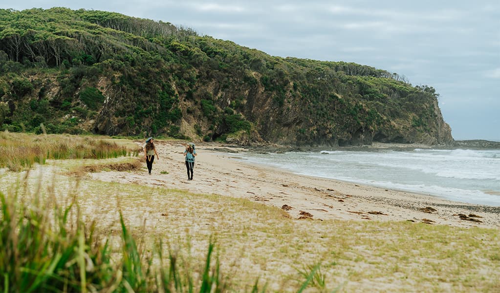2 bushwalkers walking along the sand of Oaky Beach, Murramarang National Park. Credit: Remy Brand © Remy Brand