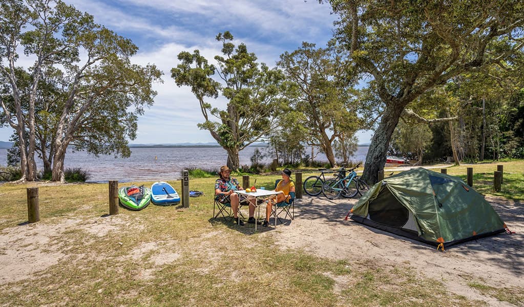 2 campers at a camping table near a tent by the water at Mungo Brush campground, Myall Lakes National Park. Credit: John Spencer/DCCEEW © DCCEEW
