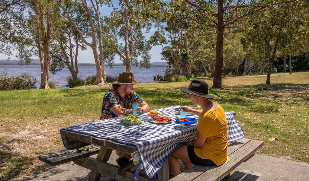 2 people having a picnic on a table by the water at Mungo Brush campground, Myall Lakes National Park. Credit: John Spencer/DCCEEW © DCCEEW