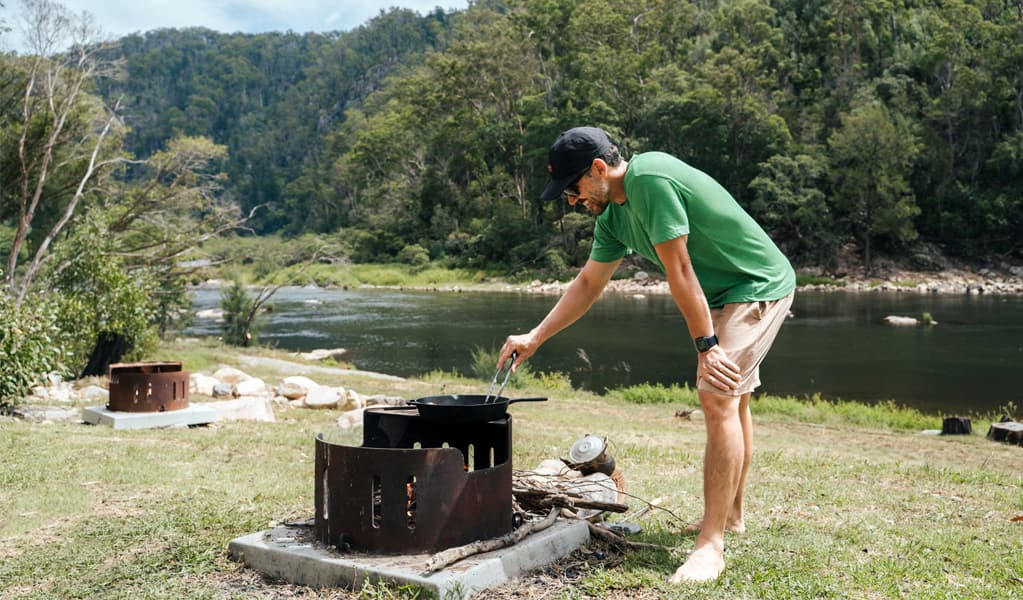 Person cooking on a fire pit at The Junction campground by Nymboida River, Nymboi-Binderay National Park. Photo: Jay Black, © DCCEEW
